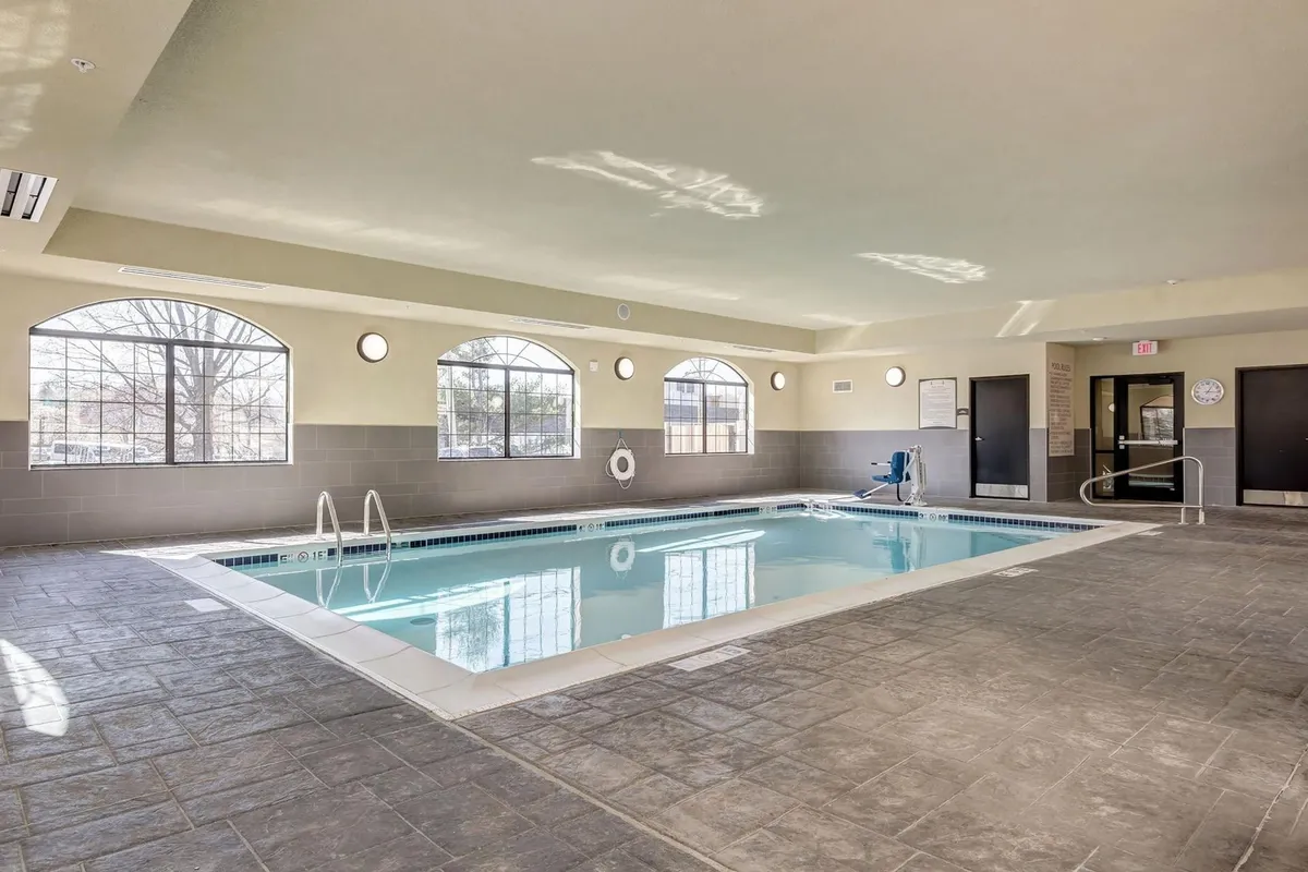 A spacious indoor swimming pool area with large windows that let in abundant natural light. Poolside seating lines the edge, inviting guests to relax after a swim. The calm atmosphere hints at both relaxation and social opportunities, perfect for families and solo travelers alike.