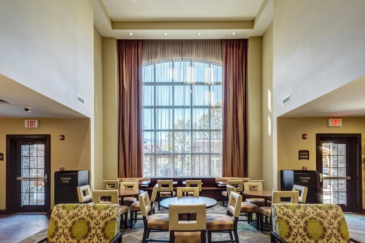 An inviting shared lounge area with round tables, upholstered chairs, and a large window draped with sheer curtains. The relaxed setting is ideal for socializing, enjoying breakfast, or unwinding during evening receptions.