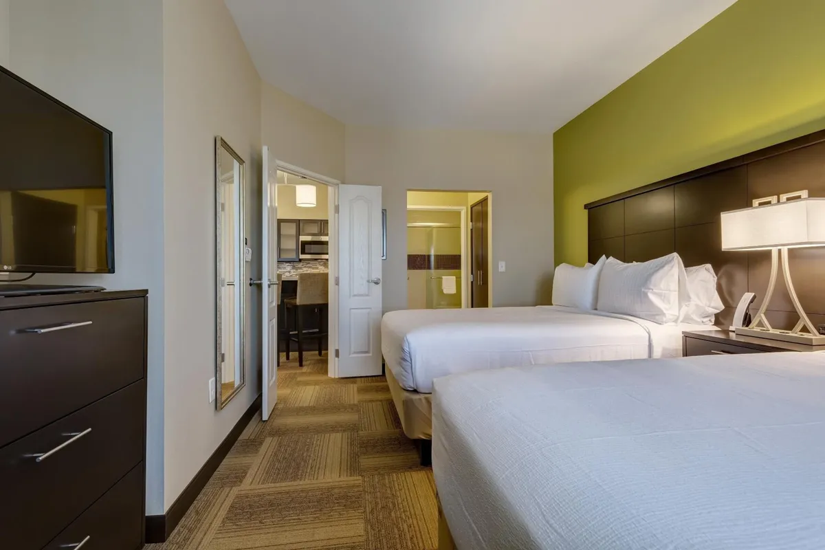 A cozy suite interior featuring two beds, a TV, a lamp, and a doorway leading to the kitchen. The setup suggests both a relaxing space for unwinding after a busy day and the convenience of in-room entertainment, making it suitable for families, colleagues, or friends. The area appears inviting and flexible, ideal for both rest and connection.