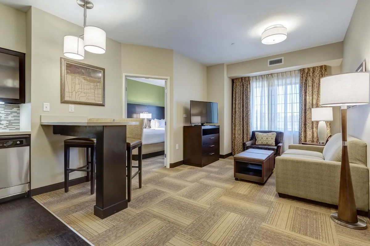 The image showcases a bright, welcoming interior of the condo’s living area. There’s a modern kitchenette with a breakfast bar, a cozy armchair, a comfortable sofa, and a large window flooding the space with natural light. The TV unit and open layout invite guests to relax, socialize, or enjoy a meal together.