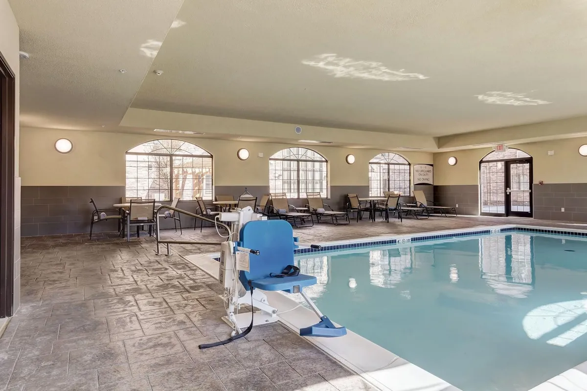 An indoor swimming pool area featuring large windows that let in ample natural light, accessibility features like a pool chair, and comfortable lounge chairs for relaxation. The tranquil environment is perfect for unwinding after a productive day.