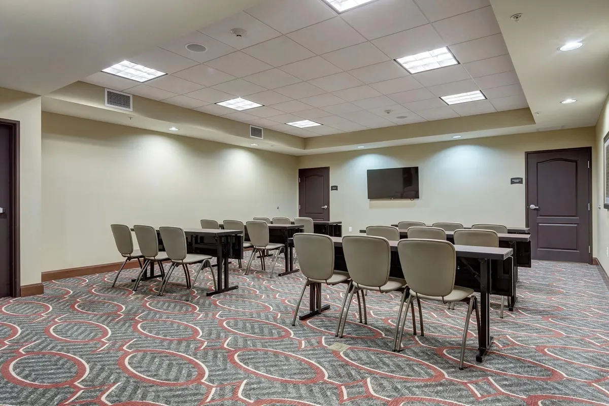 This image displays a well-lit interior space featuring tables and chairs arranged for group gatherings, with a large screen and doors visible in the background. The setting is conducive to meetings, socializing, or casual hangouts, with an inviting, collaborative atmosphere.